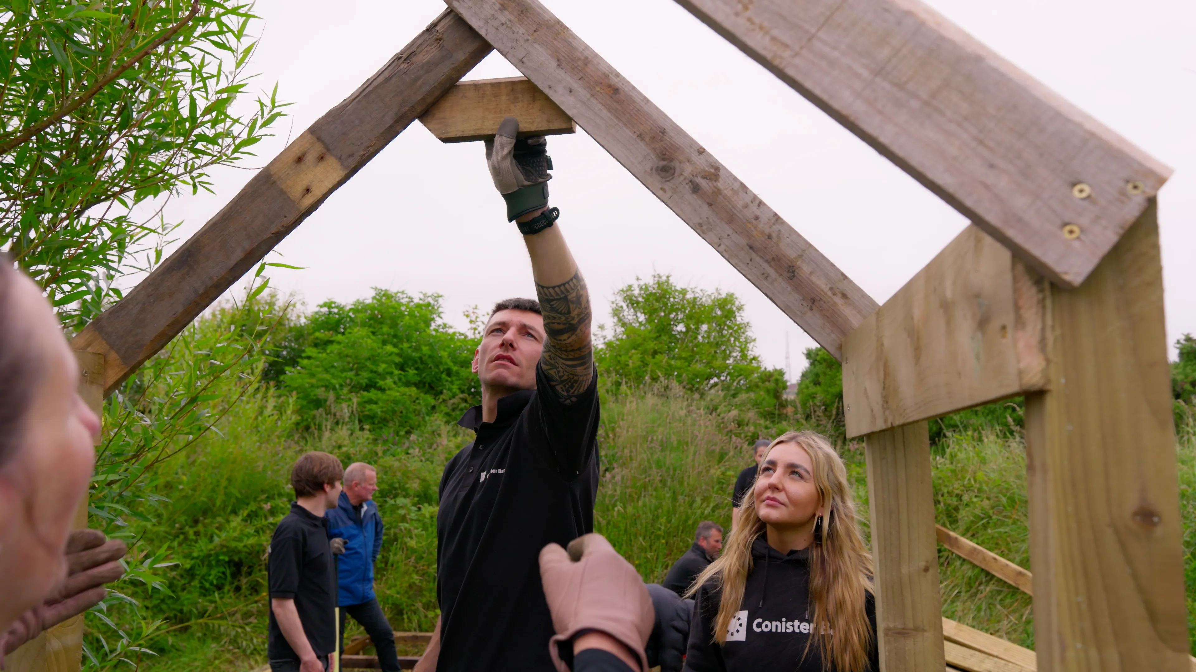A group of people work together to build a wooden structure outdoors. One person holds a beam, while others watch attentively. The scene is collaborative and focused.